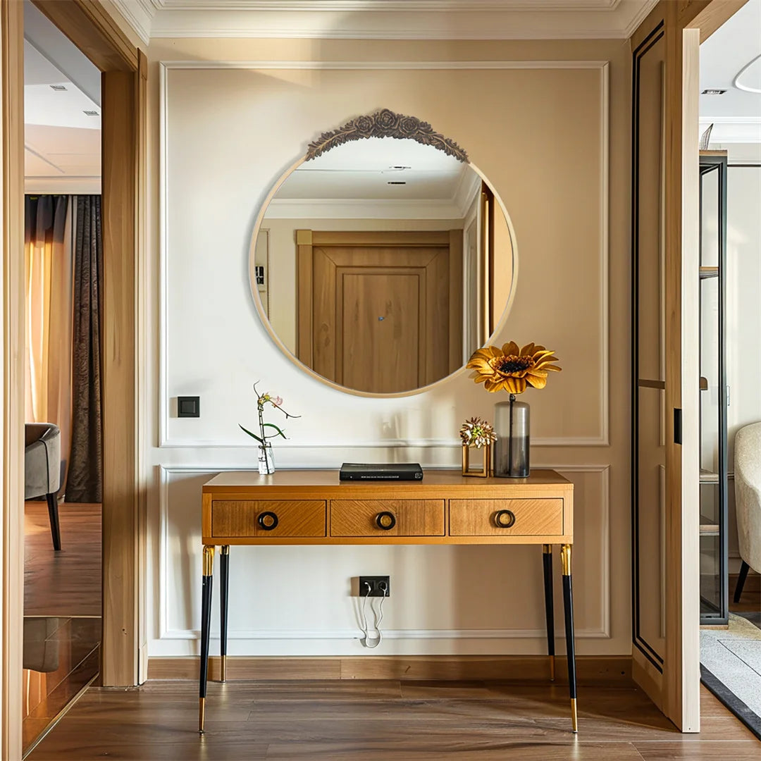 Un miroir rond doré orné de fleurs sculptées sur un meuble élégant en bois clair avec des tiroirs, placé dans un couloir lumineux avec un sol en bois.