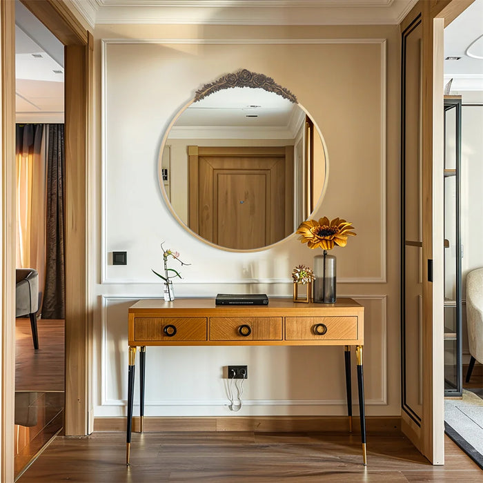 Un miroir rond doré orné de fleurs sculptées sur un meuble élégant en bois clair avec des tiroirs, placé dans un couloir lumineux avec un sol en bois.