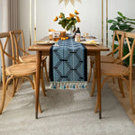 Une table en bois avec des chaises assorties, une chemin de table bleu et blanc orné de glands, un bouquet de fleurs jaunes en vase et des accessoires de décoration sur la table, offrant une scène chaleureuse et élégante dans un intérieur moderne.