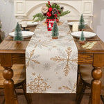 La décoration de table de Noël comprend une nappe avec des motifs de flocons de neige dorés, des figurines de sapins verts et une composition florale rouge et verte dans un vase.
