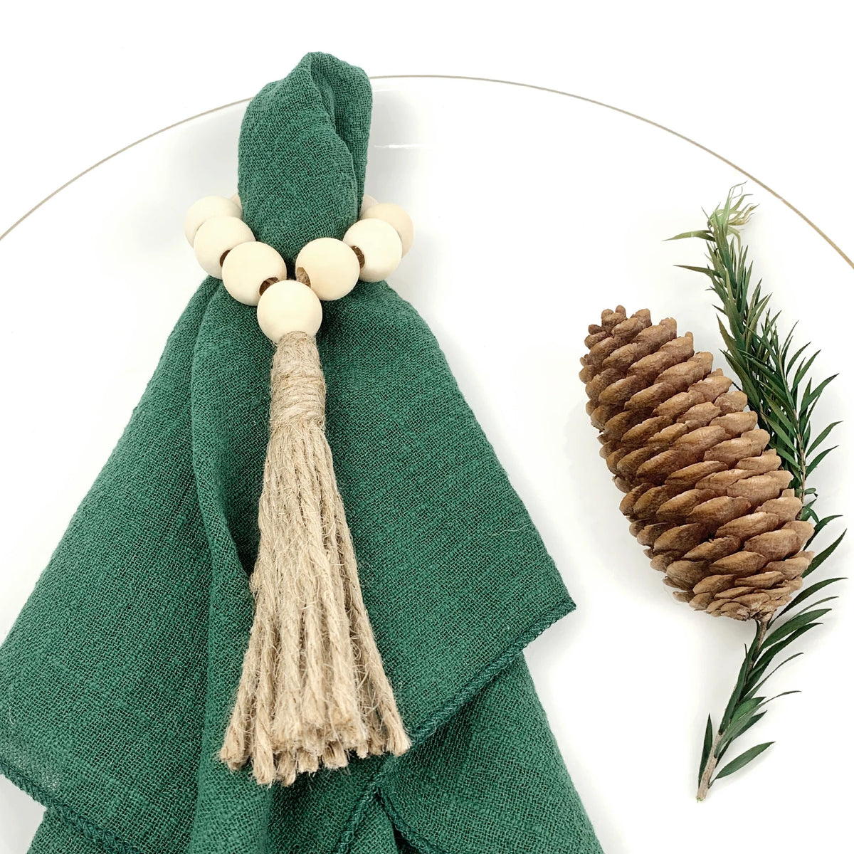 Un décor de table élégant avec une serviette verte ornée de perles de bois et d'une longue frange en corde, accompagné d'un cône de pin brun et de quelques feuilles vertes.