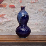 Vase en céramique bleu marine de forme élégante et classique, posé sur une surface en bois, avec un arrière-plan de briques rouges.