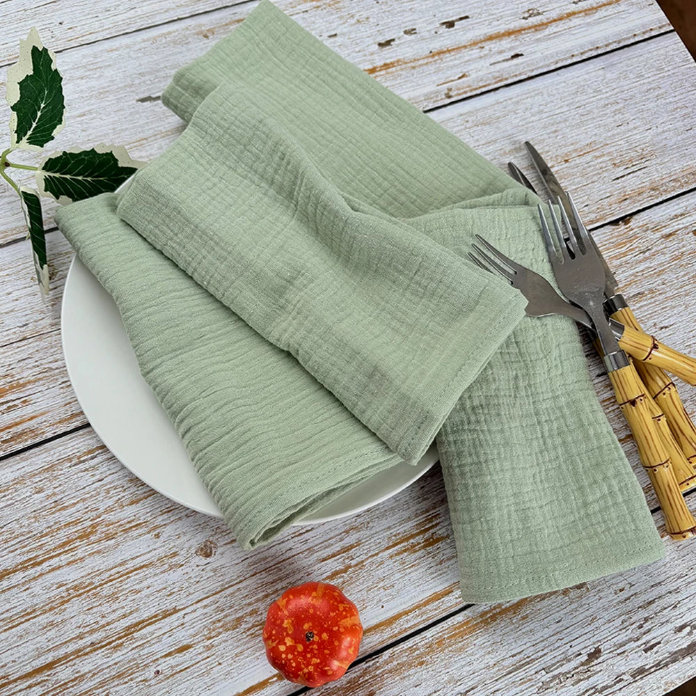 Un linge de table vert pâle, des couverts en métal et une tomate rouge sur une table en bois rustique.