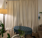 Salon moderne avec rideaux translucides beige créant une ambiance douce. Grand pouf bleu capitonné en avant-plan, entouré de plantes vertes. Lampe d'appoint à gauche offrant un éclairage chaleureux. Idéal pour un espace de détente ou de lecture.