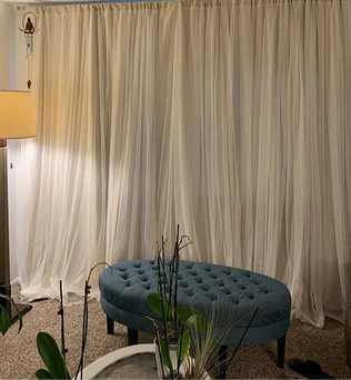Salon moderne avec rideaux translucides beige créant une ambiance douce. Grand pouf bleu capitonné en avant-plan, entouré de plantes vertes. Lampe d'appoint à gauche offrant un éclairage chaleureux. Idéal pour un espace de détente ou de lecture.