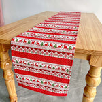 Cette image montre un chemin de table de Noël orné de motifs rouges et verts, avec des rennes et des flocons de neige répétés sur le tissu.