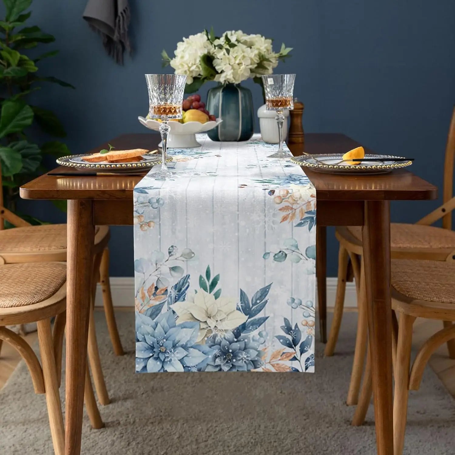 Une table dressée de manière élégante avec un chemin de table fleuri, des verres à vin et un bouquet de fleurs blanches dans un vase bleu sur un fond sombre.