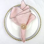 Serviette de table pliée en éventail rose pâle avec épingle décorative en métal doré en forme de fleur posée sur un plateau de service rond orné de perles dorées.