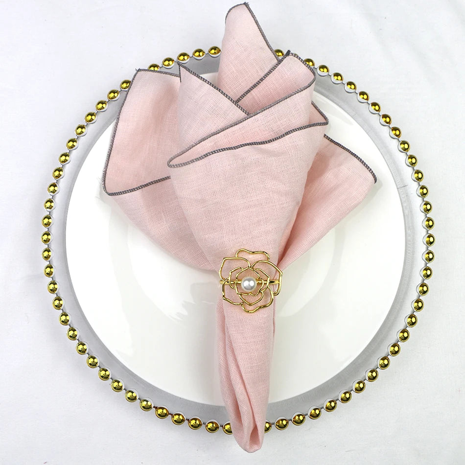 Serviette de table pliée en éventail rose pâle avec épingle décorative en métal doré en forme de fleur posée sur un plateau de service rond orné de perles dorées.