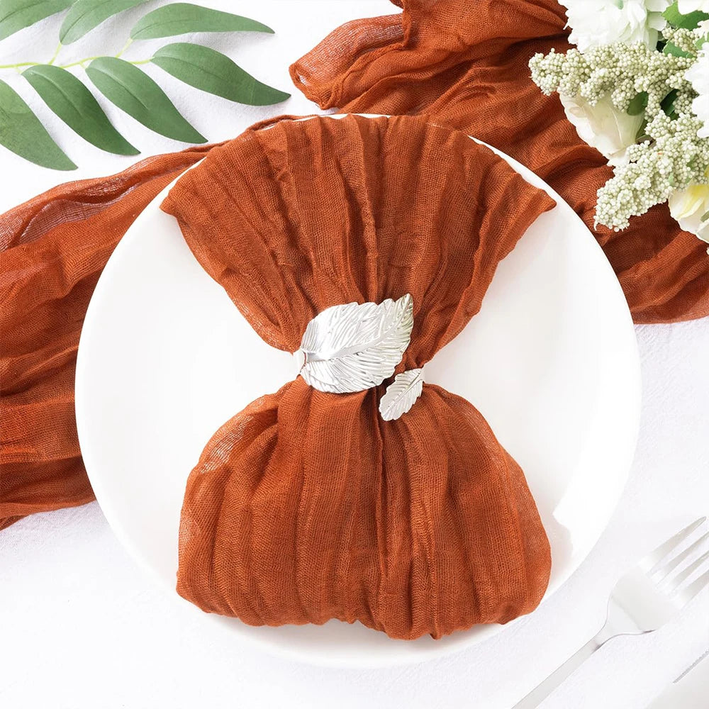 Un ravissant paquet cadeau enveloppé dans un tissu marron rougeâtre, retenu par une boucle argentée en forme de feuille, posé sur un fond blanc avec des feuilles vertes et des fleurs blanches.