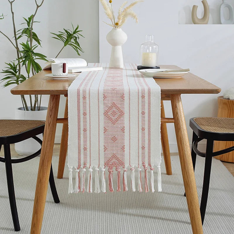 Table couverte d'un chemin de table décoratif avec des motifs géométriques et des franges, accompagné de décors végétaux et de bougies, créant une ambiance chaleureuse et épurée dans un intérieur moderne.