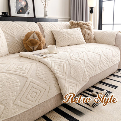 L'image montre un canapé confortable et douillet recouvert d'un plaid aux motifs géométriques beiges, accompagné de coussins décoratifs et d'un vase avec des branches, dans un décor de style rétro.