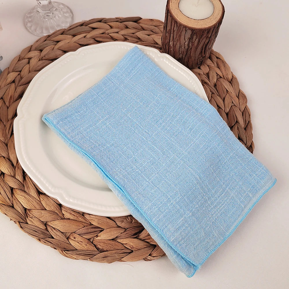 Une serviette bleu clair posée sur une assiette blanche, entourée d'un napperon en osier tressé, avec un photophore en bois à côté.