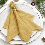 Serviette de table pliée de couleur dorée, ornée de perles, posée sur une assiette blanche entourée de fleurs.