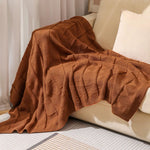 Cette image montre une couverture en tricot de couleur brune, drapée sur un canapé blanc, offrant une texture et une apparence réconfortante.