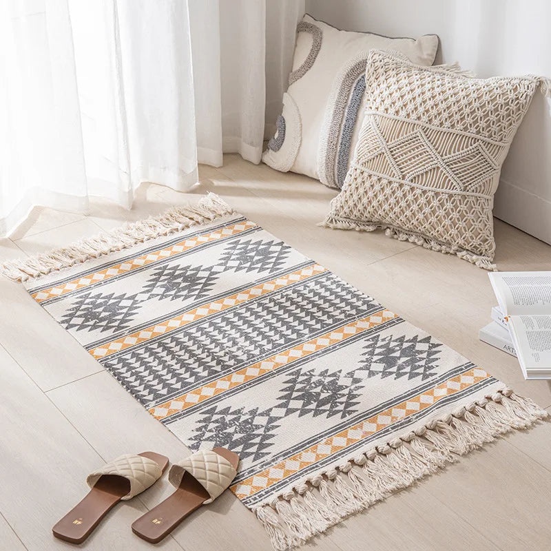Un tapis décoratif avec des motifs géométriques et des franges bohèmes, placé sur un sol clair à côté d'un coussin décoratif et de pantoufles.
