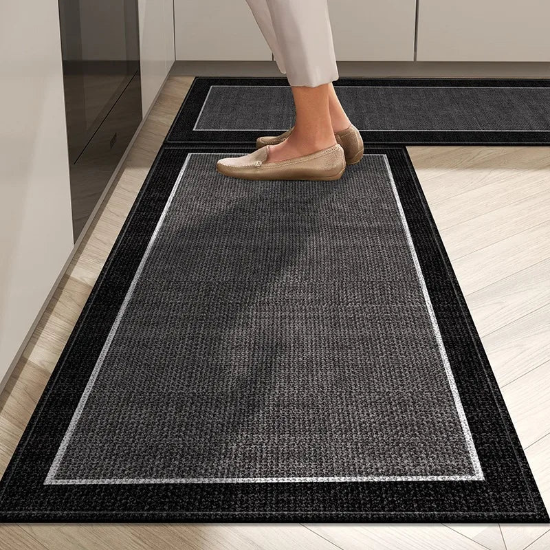 Un tapis d'entrée gris foncé avec des bordures noires, sur un sol en parquet clair, sur lequel une personne porte des chaussures beiges.