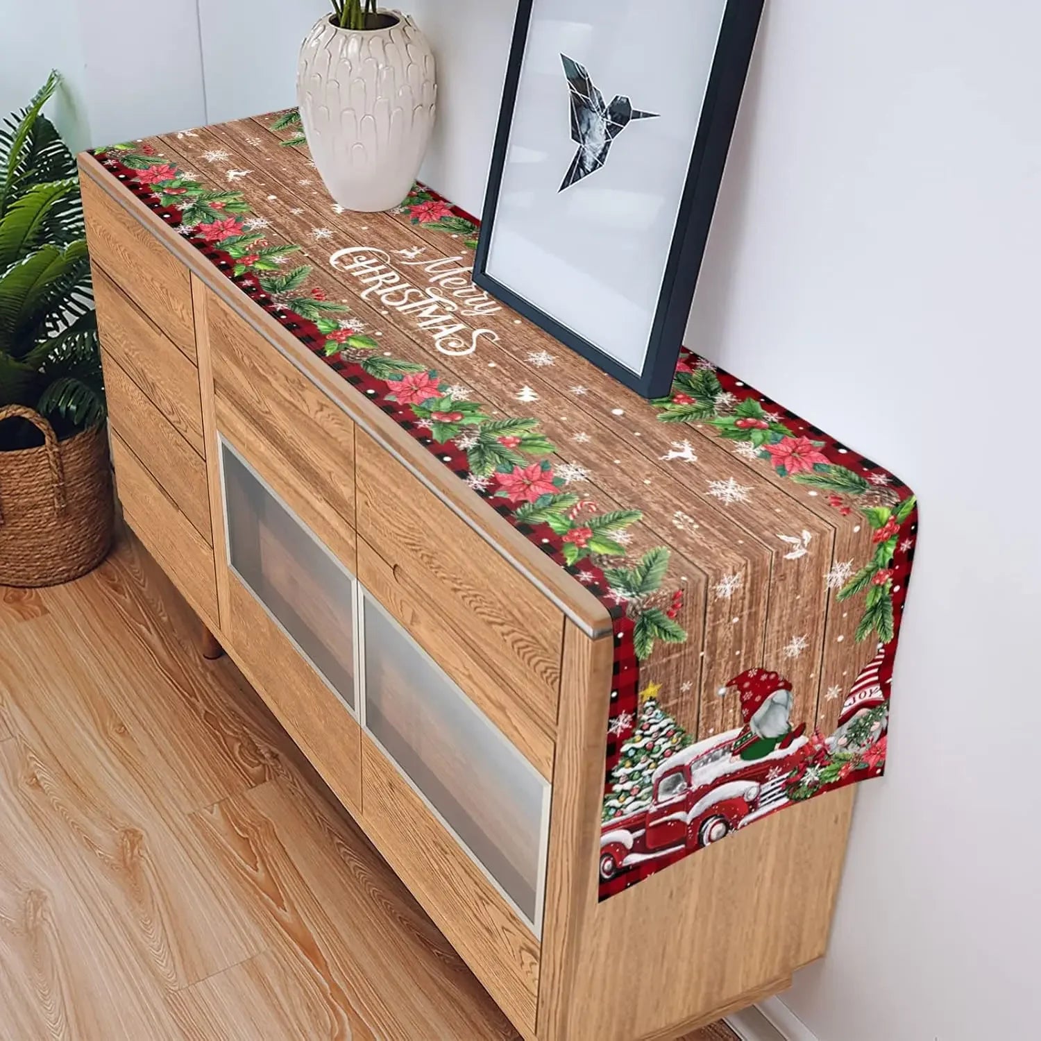 Un mobilier de salle à manger en bois avec un chemin de table aux motifs de Noël, comprenant des branches de sapin, des fleurs rouges et un camion vintage rouge.