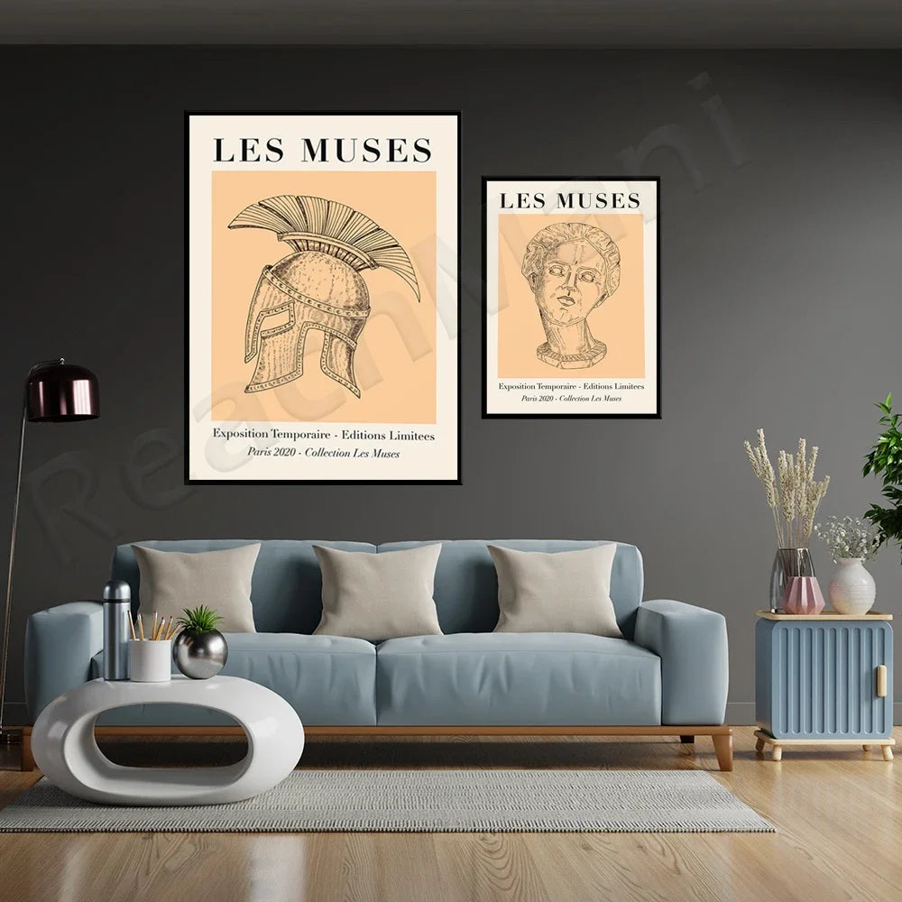 L'image montre deux affiches encadrées intitulées "Les Muses", présentant des illustrations d'un casque spartiate et d'un buste antique, exposées dans un salon contemporain décoré avec un canapé bleu gris, une table basse ronde, et des accessoires décoratifs.