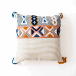 Coussin décoratif de style ethnique avec motifs géométriques colorés sur fond gris et blanc, avec des franges aux coins pour une touche bohème.
