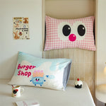Coussin en forme d'yeux avec un nez rose sur un lit, ainsi qu'un oreiller avec un personnage de dessin animé représentant une boutique de hamburgers.