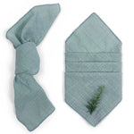 Serviette pliée verte pâle avec un ornement de branche de conifère, suggérant une ambiance apaisante et naturelle.