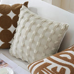 Coussin décoratif crème à motifs géométriques et pomponné, placé sur un canapé blanc avec des coussins marron, créant une ambiance cosy et chaleureuse.