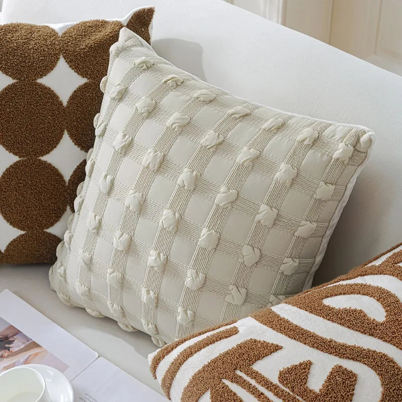 Coussin décoratif crème à motifs géométriques et pomponné, placé sur un canapé blanc avec des coussins marron, créant une ambiance cosy et chaleureuse.