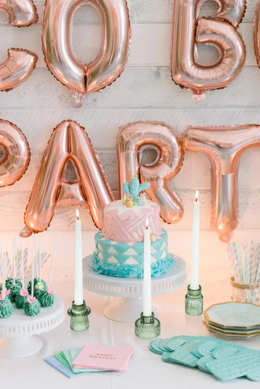 Une fête d'anniversaire rose et bleue avec des ballons dorés formant le mot "PARTY" et un gâteau décoré avec des motifs géométriques et des bougies vertes.