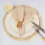 Une assiette blanche ornée d'une serviette beige enroulée autour d'un anneaux décoratif doré, posée sur un napperon tressé de couleur champagne, accompagné d'un couvert en acier.