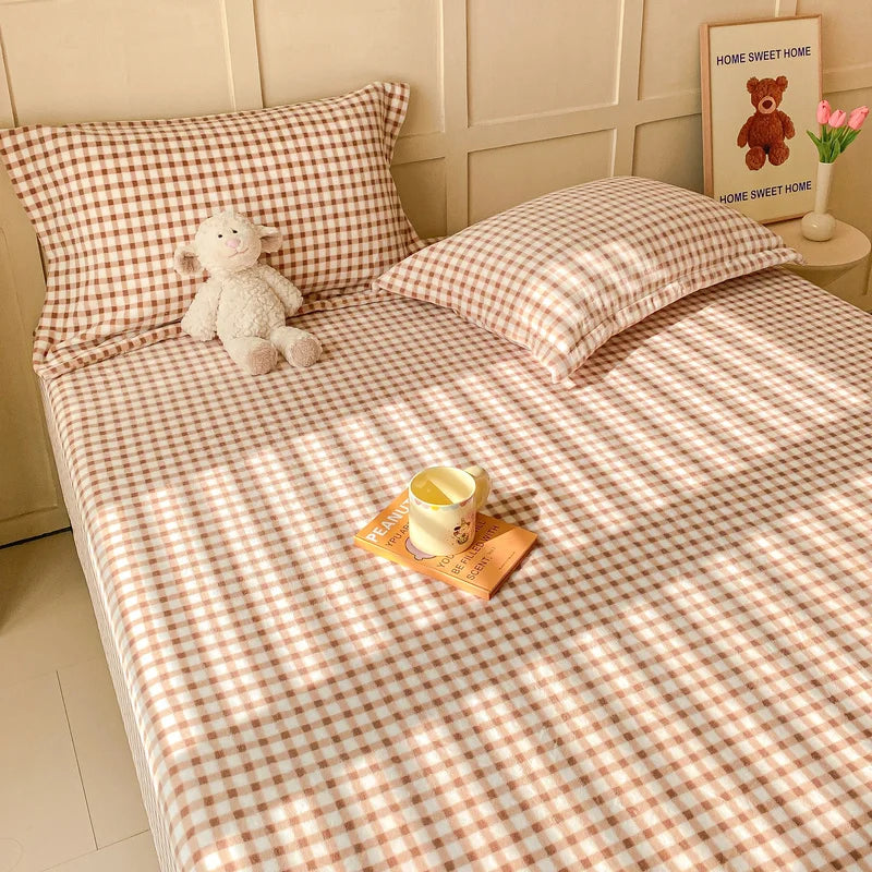 Une chambre confortable avec des draps à carreaux beiges et un ours en peluche posé sur le lit, accompagnés d'une bougie et d'une décoration murale.