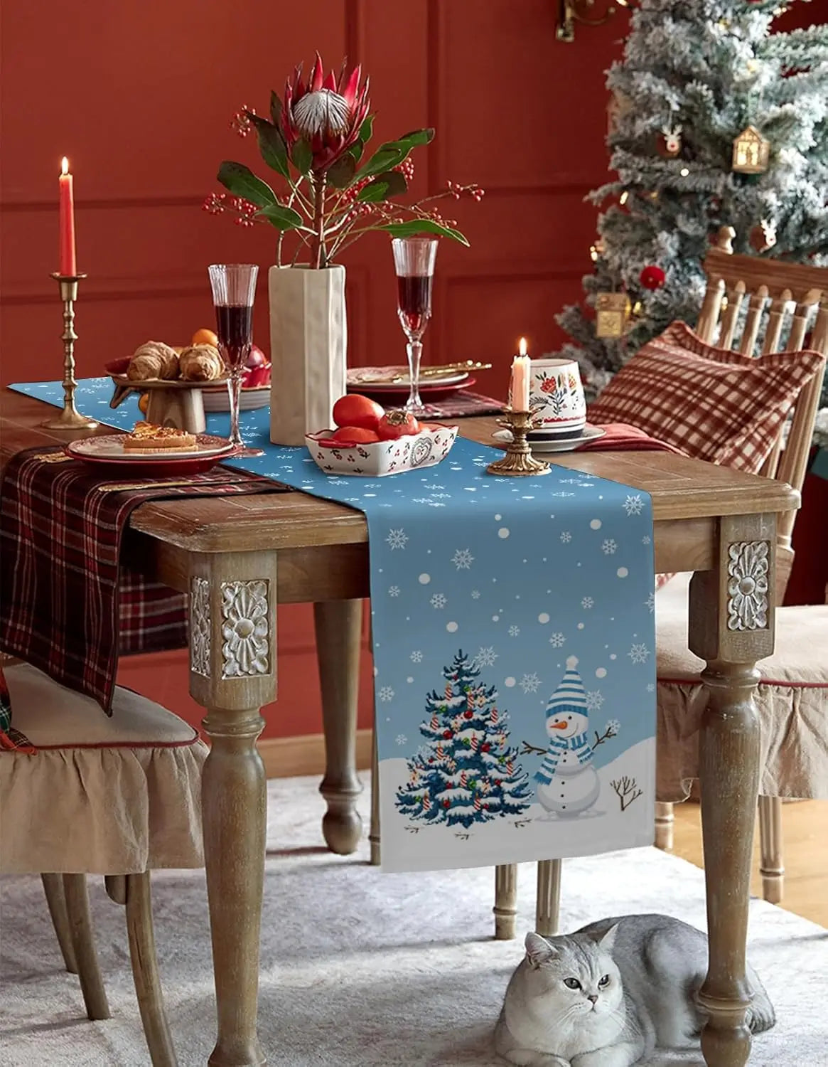 Une table de fête festive avec un arbre de Noël, des chandeliers, des verres à vin et un chat relaxant dans un décor hivernal.