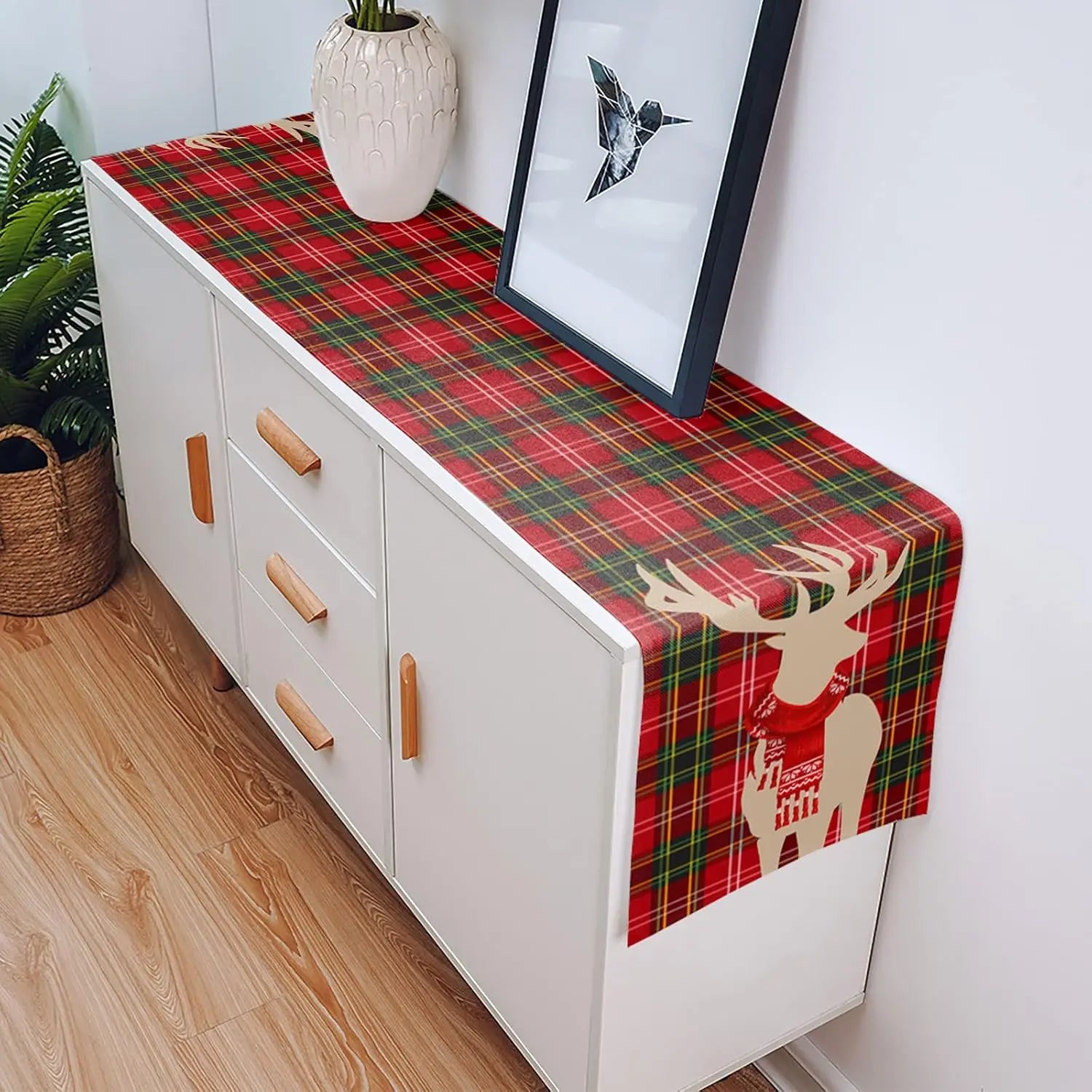 Une table en bois blanc avec un chemin de table à motif écossais et un décor de Noël, une suspension en forme d'oiseau dans un cadre noir et des plantes vertes à côté.