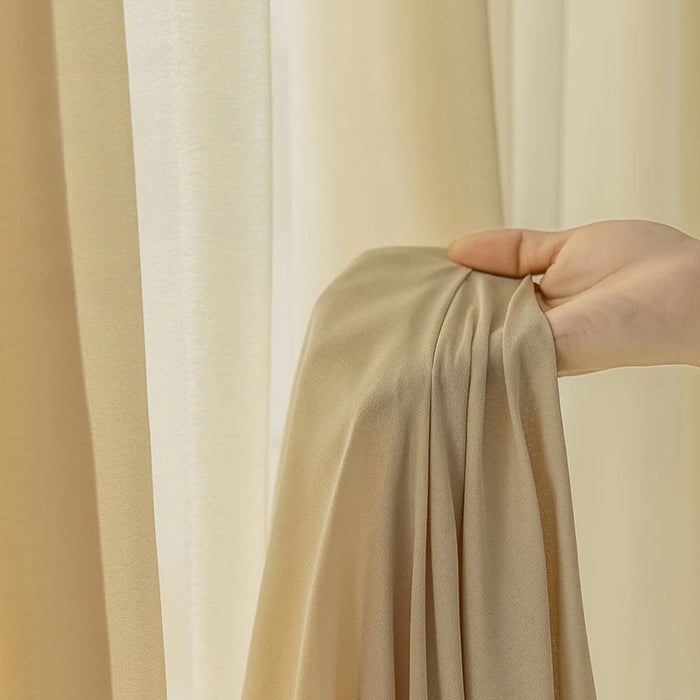 Main tenant un voilage écru fluide et élégant devant une fenêtre habillée de rideaux assortis, laissant filtrer une lumière douce et naturelle.