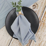 Serviette grise en tissu posée sur un plat noir circulaire avec une branche verte de plantes sur le côté, sur un fond en bois rustique.