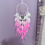 Un attrapeur de rêves décoratif rose et blanc, suspendu sur un fond violet, ornée de plumes délicates et d'un centre en forme de fleur.