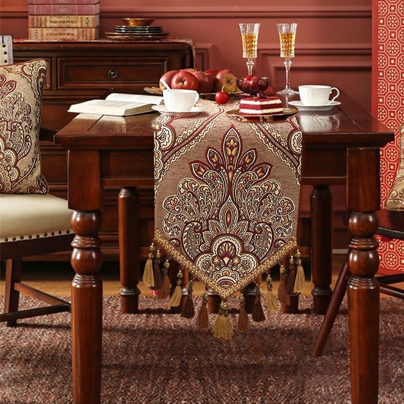 Un superbe chemin de table de style oriental, avec un design floral décoratif et des glands, sur une table de salle à manger en bois riche et chaleureuse, complétée par des assiettes, verres et autres accessoires.