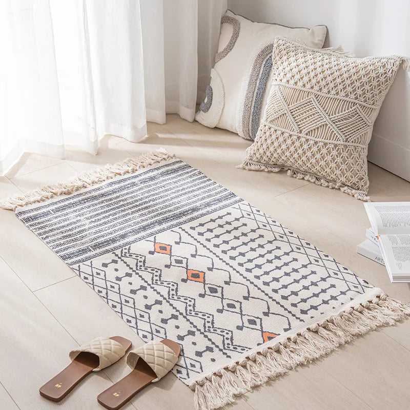 Tapis décoratif aux motifs géométriques gris et beige, orné de franges, accompagné de coussins et de sandales dans un intérieur lumineux.