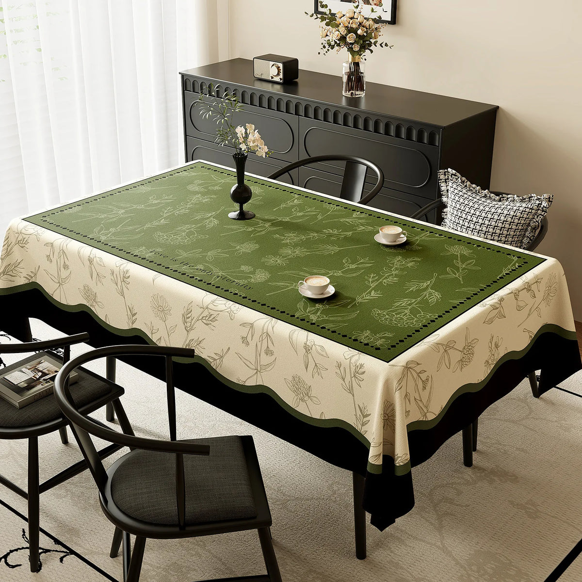 Cette image présente une belle table dressée avec une nappe imprimée de fleurs et de feuillages en vert et beige, accompagnée d'un meuble de rangement foncé orné de motifs floraux, ainsi que de quelques décorations florales posées sur la table.