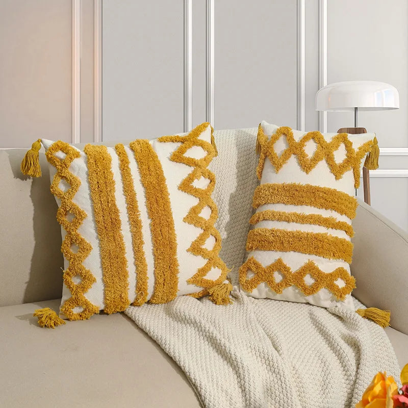 Coussin décoratif moelleux avec motifs géométriques jaunes et crème, orné de franges, posé sur un plaid en tricot dans un intérieur lumineux.