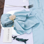 Une serviette de table en tissu bleu pâle décorée d'une broche ornée de perles et de cristaux, accompagnée d'une fourchette métallique posée à côté.