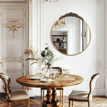 Cette image montre une salle à manger élégante avec une table ronde en bois, des chaises en bois sculptées, un miroir rond doré et des décorations florales dans un vase sur la table.