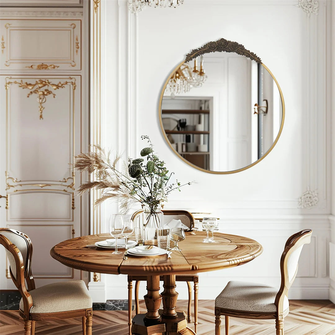 Cette image montre une salle à manger élégante avec une table ronde en bois, des chaises en bois sculptées, un miroir rond doré et des décorations florales dans un vase sur la table.
