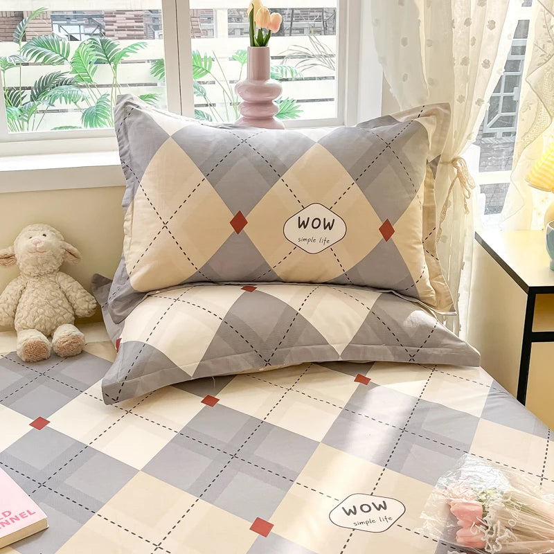 Cette image montre un ensemble de literie composé d'oreillers et d'une couverture avec un motif géométrique en gris, beige et rouge, ainsi que le texte "wow simple life" imprimé dessus. L'environnement est accueillant avec une fenêtre donnant sur des plantes et un jouet en peluche à proximité.