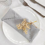 La nappe grise en tissu avec un noeud de ficelle et de feuilles dorées sur une assiette blanche évoque une atmosphère élégante et naturelle pour la table.