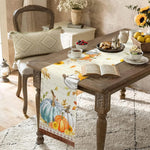 Une table de cuisine rustique avec des décorations d'automne, une serviette imprimée avec des citrouilles et des fleurs, un livre ouvert et un arrangement floral dans un vase.