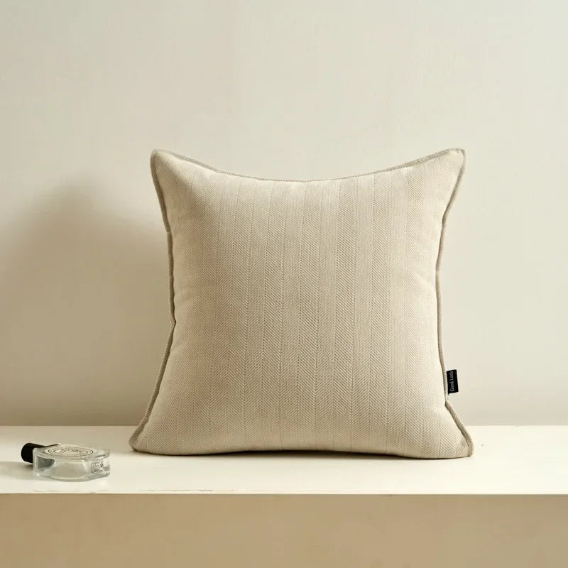 Coussin décoratif en lin beige sur une surface blanche avec un vase en verre sur le côté.