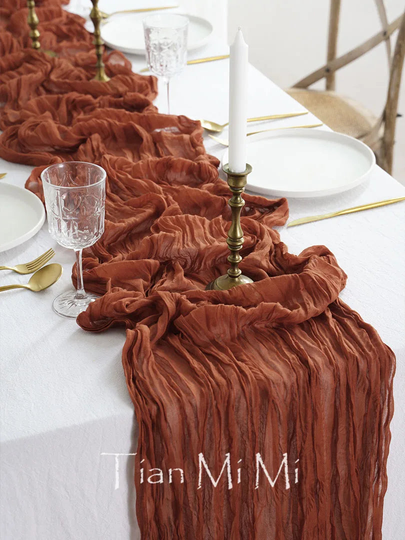 Une table de fête élégante avec des éléments décoratifs chauds et sophistiqués, incluant une nappe et une décoration froissée en tissu dans des tons rougeâtres.