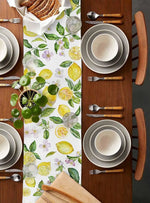 La table est joliment décorée avec des assiettes, des couverts et un chemin de table aux motifs citrons, limes et feuilles vertes, créant une atmosphère fraîche et accueillante pour un repas.