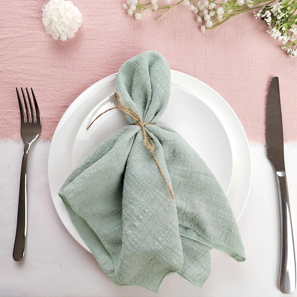 Une belle configuration de table de dîner avec un pli de serviette vert menthe décoré d'un lien en fibre naturelle, accompagné d'une fourchette et d'un couteau posés sur une assiette blanche, le tout sur un fond rose clair.